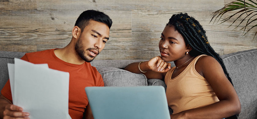 Man and woman sitting on couch looking at paperwork and computer.